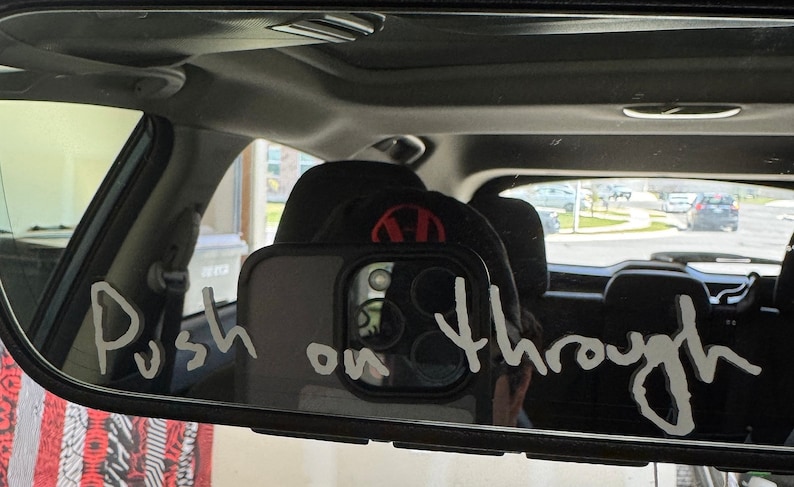 Pu&ograve; includere: Uno specchietto retrovisore in un'auto con le parole "Push on through" scritte sopra con un pennarello bianco.