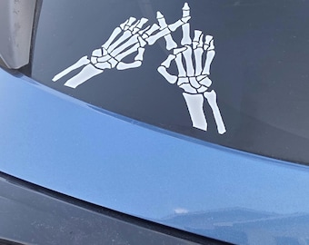 May include: White skeleton hand decal on a car window. The decal shows two hands in a peace sign gesture. The car is blue and the background is a dark color.