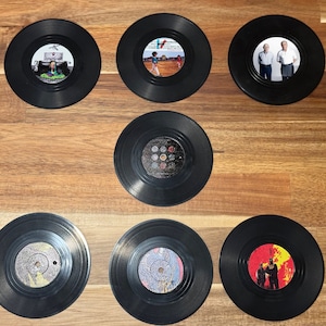 May include: Seven black vinyl record coasters with different images printed on them. The coasters are arranged in a grid pattern on a wooden surface.