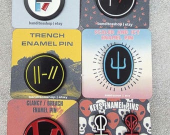 May include: A collection of six enamel pins with various designs and text. The pins feature a mix of colors including black, red, blue, and white. Each pin is on a square card with text such as "VESSEL ENAMEL PIN" and "BLURRYFACE ENAMEL PIN".