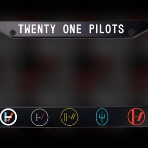 Puede incluir: Marco de matrícula negro con el texto "TWENTY ONE PILOTS" en blanco. El marco tiene seis logotipos circulares en varios colores, incluyendo rojo, azul y amarillo. El marco está diseñado para una placa de matrícula estándar.