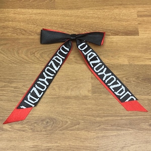 May include: A black bow with red trim and two long red and black ribbons. The ribbons have white text that reads "DJIZUXZDRO". The bow is likely an accessory for clothing or hair.