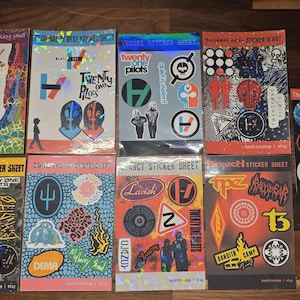 Può includere: Una collezione di fogli di adesivi colorati con vari disegni e la scritta "Twenty One Pilots". Gli adesivi includono loghi di band, simboli e grafiche, con alcuni fogli con accenti olografici. I fogli sono disposti su una superficie di legno.