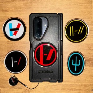 Pode incluir: Capa de telefone Otterbox preta com um logotipo vermelho e preto, rodeada por seis pinos redondos com vários desenhos em preto, vermelho, azul e amarelo. Um pino tem um pequeno cordão preto preso.