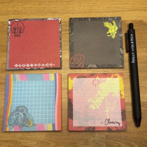 Puede incluir: Cuatro blocs de notas cuadrados con diseños únicos y un bolígrafo negro. Los blocs de notas presentan una calavera roja, un águila amarilla, un dragón azul y una cuadrícula colorida. El bolígrafo dice "ADDICT WITH A PEN".