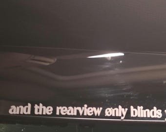 May include: A black rearview mirror with the text "and the rearview only blinds you" in white. The mirror is rectangular with rounded corners and is mounted inside a vehicle.