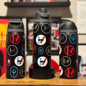 May include: Three black stainless steel water bottles with a variety of colorful logos and symbols. The logos include a trident, a circle with a slash through it, and a circle with a horizontal line through it.