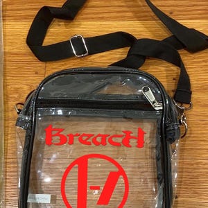 May include: Clear crossbody bag with a black strap and zipper closure. The bag features the word "Breach" in red, along with a red circle with a stylized "17" design. The bag is made of clear plastic.