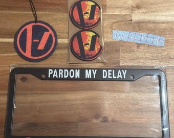 May include: A black license plate frame with the text "Pardon My Delay I'm Navigating" and a red and black logo. The frame is surrounded by a clear plastic cover. Two round coasters with a red and black logo are included. The coasters are made of a soft material and have a textured surface.