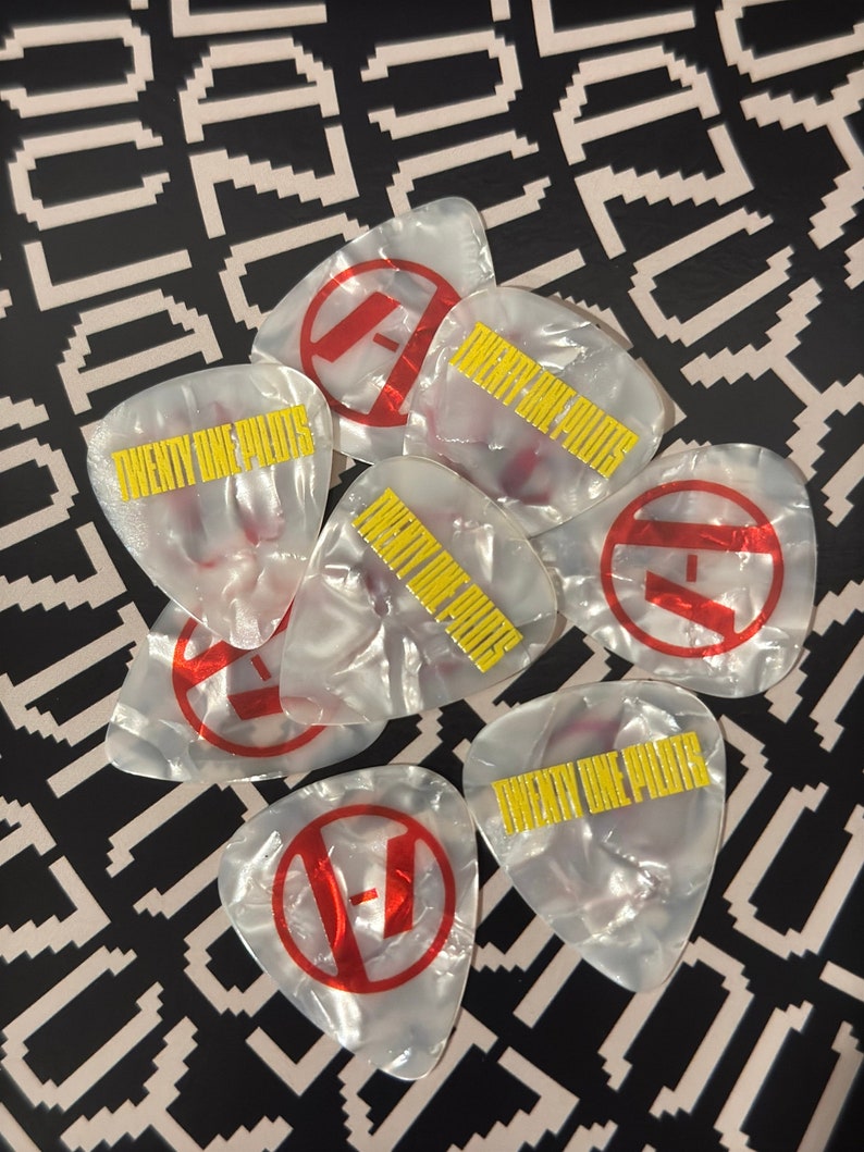 May include: Seven white guitar picks with a red circle and the letter "F" in white inside the circle. The text "TWENTY ONE PILOTS" is printed in yellow on four of the picks.