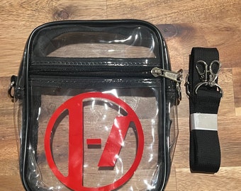 Può includere: Borsa a tracolla trasparente in plastica con cerniera nera e tracolla regolabile nera. La borsa ha un cerchio rosso con le lettere "I-7" all'interno.