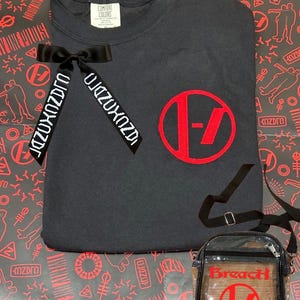 May include: A black t-shirt with a red logo and a black ribbon with white text. A clear crossbody bag with a black trim and a red logo is also included. The background features a red and black pattern.