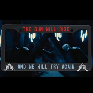 May include: Black license plate frame with the text "THE SUN WILL RISE" in red and "AND WE WILL TRY AGAIN" in white. The frame has a dark background with a blurred image.