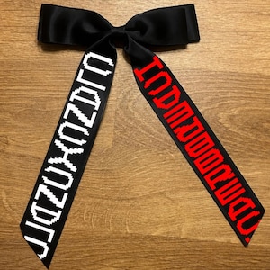 May include: A black satin bow tie with two long, decorative tails. One tail has white pixelated text, and the other has red pixelated text. The bow tie is displayed on a wooden surface.