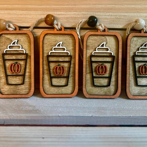 May include: Four wooden fall-themed ornaments. Each ornament features a coffee cup design with a pumpkin and whipped cream. The ornaments have a rectangular shape with a wooden frame and a string for hanging. The color scheme includes orange, brown, black, and white.