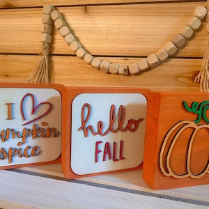 May include: Three wooden blocks with fall-themed designs. One block says "I heart pumpkin spice", another says "hello FALL", and the last features a pumpkin outline. A wooden bead garland hangs above.
