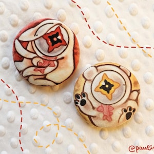 May include: Two round, fabric-covered buttons with cartoon illustrations. One button features a red and white design with a central starburst. The other button has a tan and brown design with paw prints and a starburst. The background is white with dotted texture.