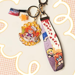 May include: A gold keychain with a purple and white strap that says "take it home!" and features cartoon characters. The keychain also has a clear charm with a cartoon character holding a bowl and a star charm.