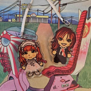 Könnte beinhalten: Zwei Acryl-Anhänger mit Illustrationen von Anime-Charakteren, die an einer weißen Kordel hängen. Der Hintergrund zeigt einen rosa Roller mit japanischem Design vor einer Straßenszene.