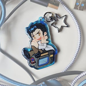 May include: A blue acrylic keychain featuring a cartoon character with black hair and a white lab coat, sitting on a retro television. The keychain has a silver star charm and a silver keyring. The background includes USB cables.