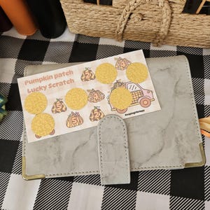 May include: A gray marble-patterned binder with a snap closure sits beneath a rectangular Pumpkin patch Lucky Scratch card. The card features illustrations of pumpkins with numbers and a truck, with five circular gold scratch-off stickers applied.