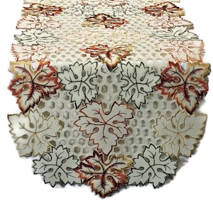 May include: White table runner with an embroidered design of autumn leaves in shades of green, orange, and brown. The leaves are arranged in a repeating pattern and have a delicate, lace-like appearance.