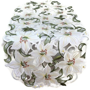 Op de afbeelding: Witte geborduurde tafelloper met een delicaat bloemenontwerp met lelies en bladeren. De loper heeft een gegolfde rand en is gemaakt van een lichtgewicht stof.