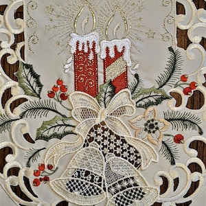 Puede incluir: Caminero de mesa blanco bordado con un tema navide&ntilde;o. El dise&ntilde;o presenta dos velas rojas con detalles dorados, un lazo, dos campanas y bayas de acebo.