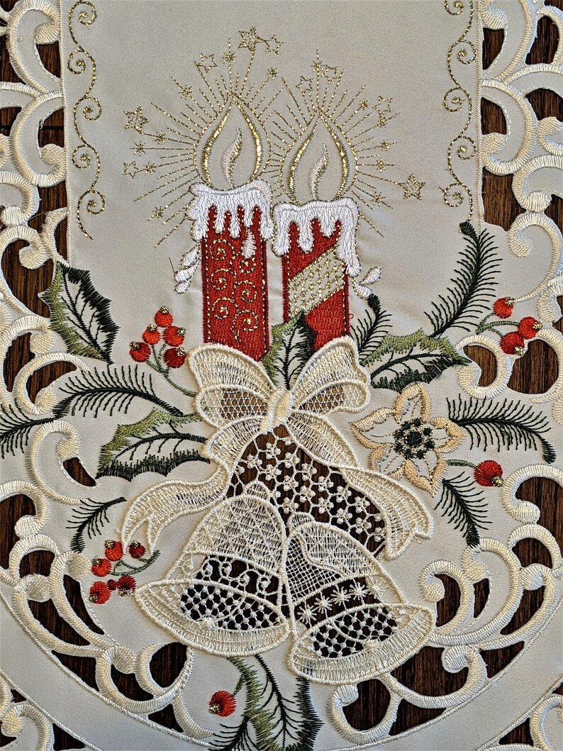 Puede incluir: Caminero de mesa blanco bordado con un tema navide&ntilde;o. El dise&ntilde;o presenta dos velas rojas con detalles dorados, un lazo, dos campanas y bayas de acebo.