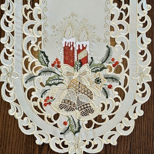 Puede incluir: Un camino de mesa blanco bordado con un tema navide&ntilde;o. El dise&ntilde;o presenta dos velas rojas con detalles dorados, un lazo y campanas. El camino de mesa tiene un borde festoneado.