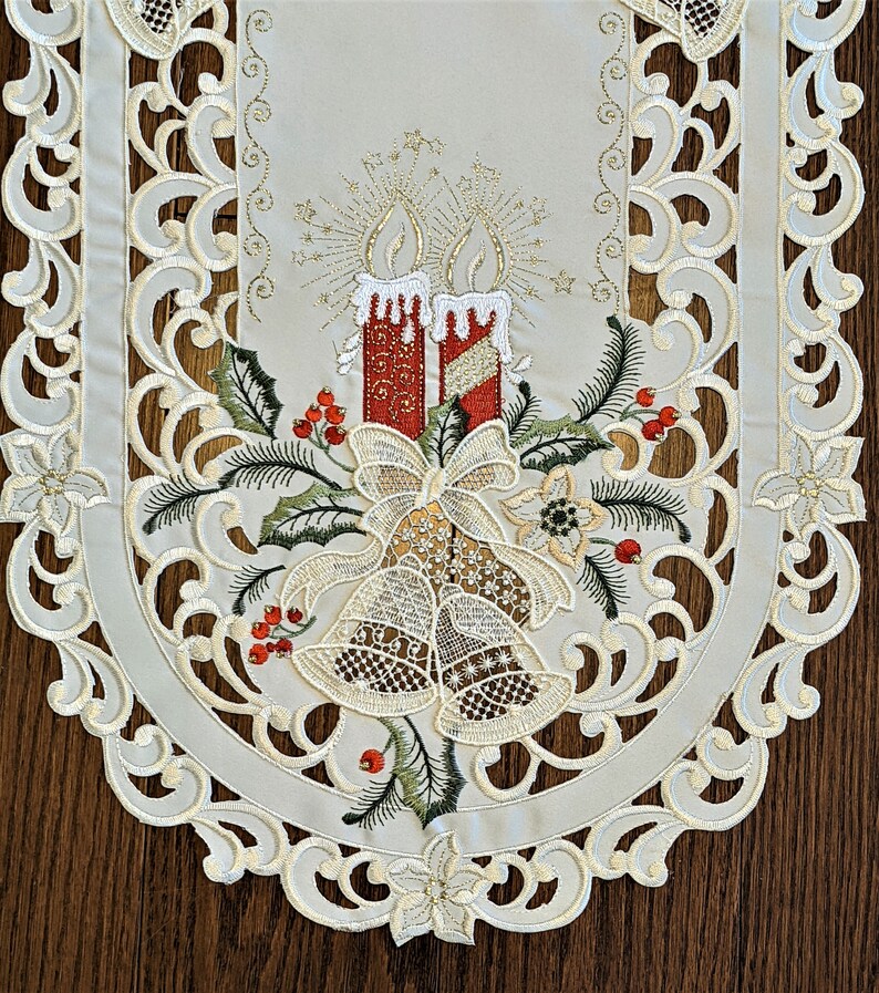 Puede incluir: Un camino de mesa blanco bordado con un tema navide&ntilde;o. El dise&ntilde;o presenta dos velas rojas con detalles dorados, un lazo y campanas. El camino de mesa tiene un borde festoneado.