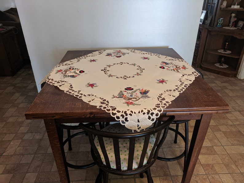 K&ouml;nnte beinhalten: Eine wei&szlig;e Tischdecke mit einer Spitzenbord&uuml;re und gestickten Winterfest-Motiven. Die Tischdecke liegt auf einem Holztisch mit zwei schwarzen St&uuml;hlen.