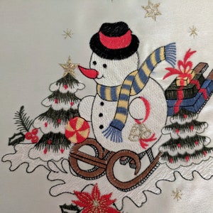 K&ouml;nnte beinhalten: Wei&szlig;e bestickte Tischdecke mit einem Schneemann-Motiv. Der Schneemann tr&auml;gt einen schwarzen Hut mit rotem Band, einen blau-gelb gestreiften Schal und h&auml;lt einen Zuckerstock in der Hand. Der Schneemann f&auml;hrt auf einem Schlitten mit einer rot-blauen Geschenkbox oben drauf.
