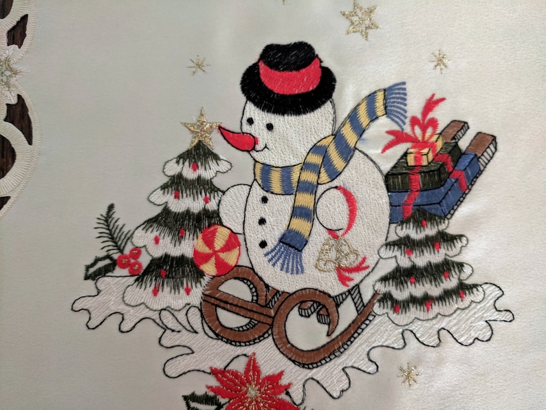 K&ouml;nnte beinhalten: Wei&szlig;e bestickte Tischdecke mit einem Schneemann-Motiv. Der Schneemann tr&auml;gt einen schwarzen Hut mit rotem Band, einen blau-gelb gestreiften Schal und h&auml;lt einen Zuckerstock in der Hand. Der Schneemann f&auml;hrt auf einem Schlitten mit einer rot-blauen Geschenkbox oben drauf.