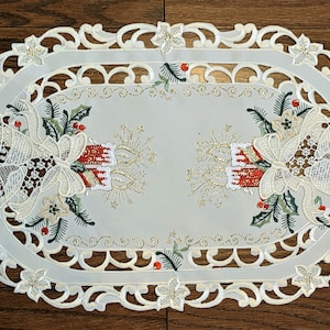 Puede incluir: Caminero de mesa ovalado blanco con un borde de encaje y dise&ntilde;os navide&ntilde;os bordados. El dise&ntilde;o presenta campanas, acebo y velas con detalles de purpurina dorada.