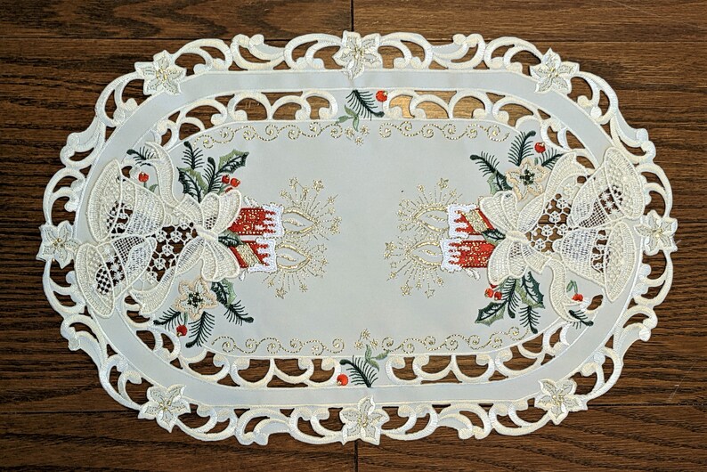 Puede incluir: Caminero de mesa ovalado blanco con un borde de encaje y dise&ntilde;os navide&ntilde;os bordados. El dise&ntilde;o presenta campanas, acebo y velas con detalles de purpurina dorada.