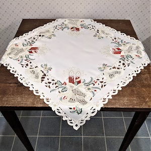 Puede incluir: Mantel blanco cuadrado con un delicado borde de encaje y motivos navide&ntilde;os bordados. El dise&ntilde;o presenta velas rojas y verdes, campanas y hojas de acebo.
