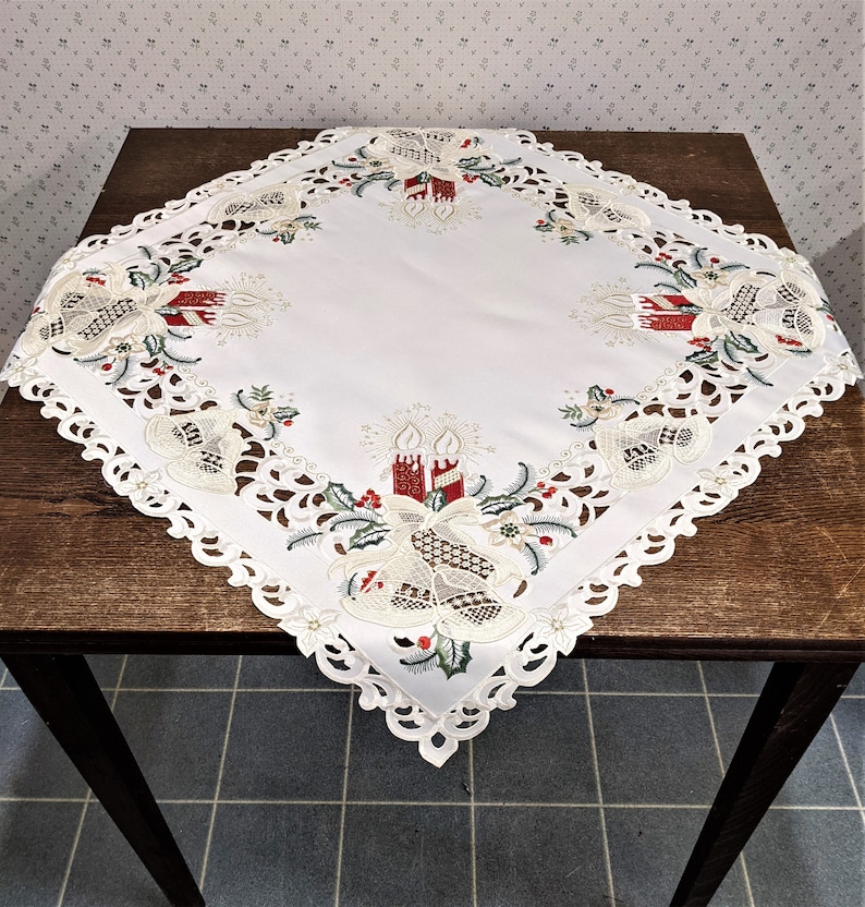 Puede incluir: Mantel blanco cuadrado con un delicado borde de encaje y motivos navide&ntilde;os bordados. El dise&ntilde;o presenta velas rojas y verdes, campanas y hojas de acebo.