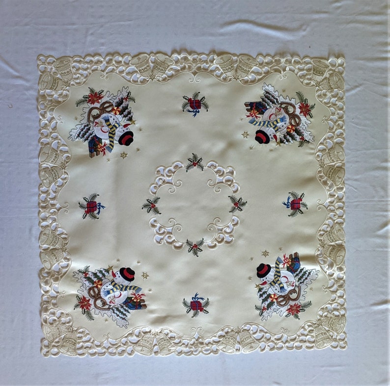 K&ouml;nnte beinhalten: Eine wei&szlig;e Tischdecke mit einer zarten Spitzenbord&uuml;re und gestickten Schneemann-Motiven. Die Tischdecke zeigt ein zentrales Design eines Schneemanns mit einem roten Schal und einem gr&uuml;nen Kranz. Die Tischdecke ist perfekt f&uuml;r ein Weihnachtsessen oder eine Feiertagsfeier.