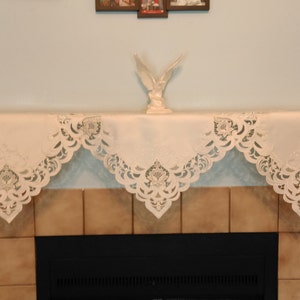 May include: Cream-colored fireplace mantel scarf with intricate cut-out floral and scroll designs. The fabric drapes over the mantel in a scalloped pattern. A white decorative bird sculpture sits on the mantel.
