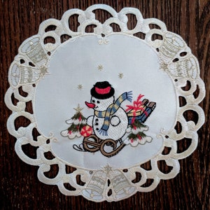 K&ouml;nnte beinhalten: Wei&szlig;e Spitze mit einem wei&szlig;en gestickten Schneemann-Design. Der Schneemann tr&auml;gt einen rot-wei&szlig;en Hut und einen gelb-blauen Schal. Der Schneemann h&auml;lt ein rot-wei&szlig;es Geschenk und zieht einen Schlitten mit einem rot-wei&szlig;en Geschenk darauf.