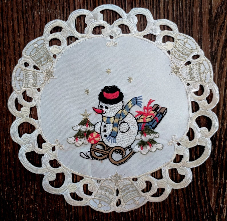K&ouml;nnte beinhalten: Wei&szlig;e Spitze mit einem wei&szlig;en gestickten Schneemann-Design. Der Schneemann tr&auml;gt einen rot-wei&szlig;en Hut und einen gelb-blauen Schal. Der Schneemann h&auml;lt ein rot-wei&szlig;es Geschenk und zieht einen Schlitten mit einem rot-wei&szlig;en Geschenk darauf.