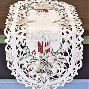 Puede incluir: Caminero de mesa ovalado blanco con un dise&ntilde;o bordado de velas rojas, campanas doradas y acebo verde. El dise&ntilde;o est&aacute; rodeado por un borde de encaje blanco.