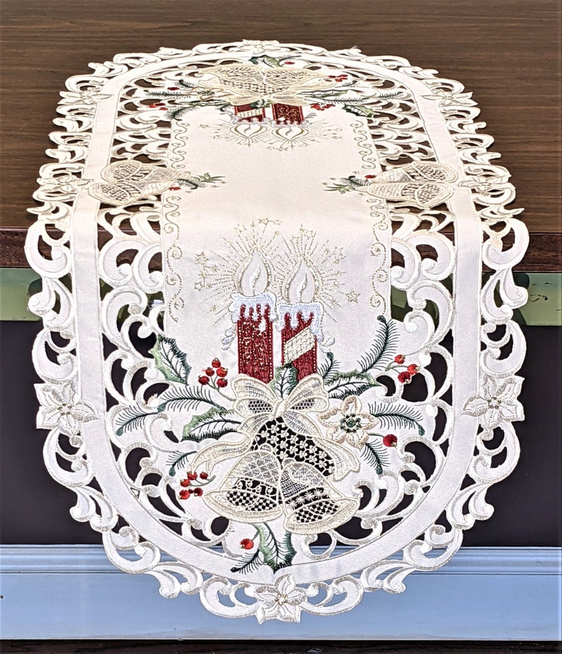 Puede incluir: Caminero de mesa ovalado blanco con un dise&ntilde;o bordado de velas rojas, campanas doradas y acebo verde. El dise&ntilde;o est&aacute; rodeado por un borde de encaje blanco.