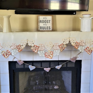 May include: A white fireplace mantel with a cream-coloured lace tablecloth featuring a fall leaf design. A sign reading "My Roost My Rules" is on the mantel. A string of fabric hearts hangs below the mantel.