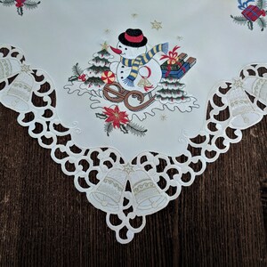 K&ouml;nnte beinhalten: Ein wei&szlig;er Tischl&auml;ufer mit einer wei&szlig;en Spitzenbord&uuml;re. In der Mitte des L&auml;ufers befindet sich ein gesticktes Design eines Schneemanns, der einen Schlitten mit Geschenken zieht. Der Schneemann tr&auml;gt einen schwarzen Hut und einen rot-wei&szlig;en Schal. Das Design beinhaltet auch rote und gr&uuml;ne Stechpalmen und Glocken.