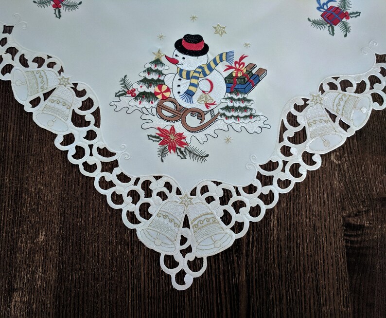 K&ouml;nnte beinhalten: Ein wei&szlig;er Tischl&auml;ufer mit einer wei&szlig;en Spitzenbord&uuml;re. In der Mitte des L&auml;ufers befindet sich ein gesticktes Design eines Schneemanns, der einen Schlitten mit Geschenken zieht. Der Schneemann tr&auml;gt einen schwarzen Hut und einen rot-wei&szlig;en Schal. Das Design beinhaltet auch rote und gr&uuml;ne Stechpalmen und Glocken.