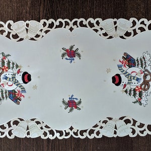 K&ouml;nnte beinhalten: Ein wei&szlig;er Tischl&auml;ufer mit einer Spitzenbord&uuml;re und gestickten Schneem&auml;nnern mit Weihnachtsb&auml;umen und Geschenken. Der L&auml;ufer hat einen gewellten Rand.