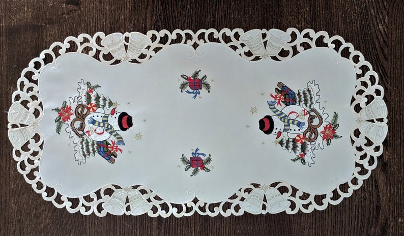 K&ouml;nnte beinhalten: Ein wei&szlig;er Tischl&auml;ufer mit einer Spitzenbord&uuml;re und gestickten Schneem&auml;nnern mit Weihnachtsb&auml;umen und Geschenken. Der L&auml;ufer hat einen gewellten Rand.