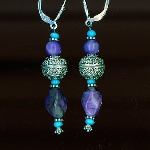 May include: A pair of drop earrings with silver lever-back fastenings. Each earring features purple and turquoise beads, including ornate silver filigree beads. The earrings are set against a dark background.